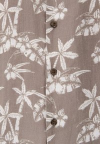 Camisa de lino ligera con un fondo beige y patrones florales y de hojas tropicales en blanco, cerrada con botones marrones.