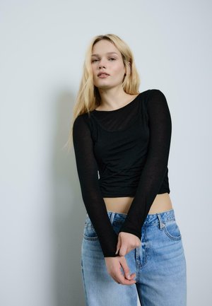 Ung kvinde med langt blondt hår iført en sort langærmet top og lyseblå jeans, stående mod en ensfarvet lys baggrund.
