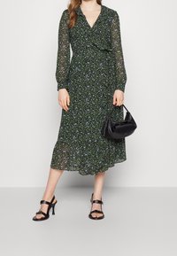 Mujer vistiendo un vestido midi de flores verdes de manga larga, sandalias de tacón negras, sosteniendo un pequeño bolso negro frente a un fondo liso.