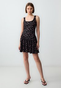 Zwarte mouwloze jurk met een wit en roze bloemenpatroon, een getailleerde bodice, een uitlopende rok en een gerimpelde zoom, gemaakt van lichte stof.