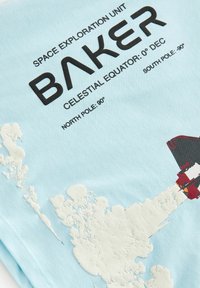 Šviesiai mėlynas audinio marškinėlis su juodu tekstu. Dizainas apima raudoną raketą su baltais debesimis. Ekrano spausdinti grafikos elementai.