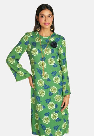 Grünes, blumig gemustertes Kleid mit langen Ärmeln, das weiße und blaue Rosen sowie blaue Blätter zeigt. Enthält eine schwarze Blumenakzent und goldenes Schmuck.