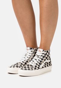 Wysokie sneakersy w czarno-białą szachownicę. Górna część z materiału, białe sznurowadła i gumowa podeszwa. Minimalistyczne logo na języku.