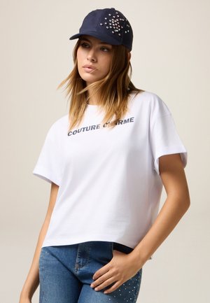 Witte katoenen T-shirt met "COUTURE CHERME" in donkergrijs gedrukt. Gepaarde met een marineblauwe pet met rhinestone versieringen.
