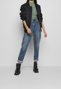 Chaqueta bomber negra sobre una camiseta de canalé verde, combinada con jeans de lavado claro y botas negras de tobillo con hebillas, en un fondo neutro.
