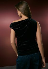 Top din catifea neagră cu design off-the-shoulder, textură căptușită și detalii strânse la talie, asociat cu blugi. Fundal în nuanță de burgund.