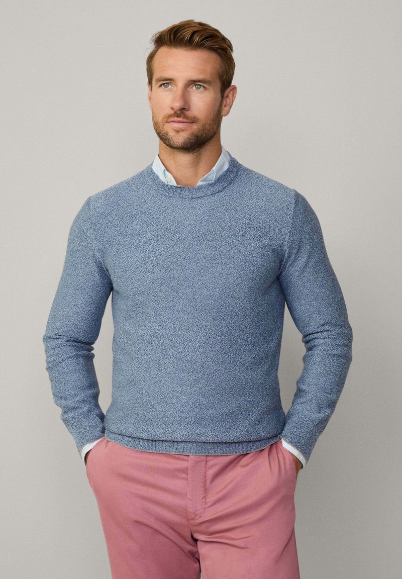 Blauer strukturierten Strickpullover mit Rundhalsausschnitt und langen Ärmeln, kombiniert mit hellrosa Hosen, vor einem neutralen Hintergrund stehend.
