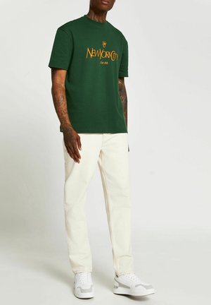 Grünes Baumwoll-T-Shirt mit dem grafischen Aufdruck "NEWYORKCITY Est 88" in Orange, kombiniert mit off-weißen Jeanshosen und weißen Sneakers.