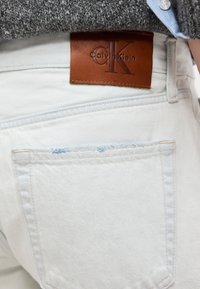 Άσπρο τζιν denim με καφέ δερμάτινο λογότυπο Calvin Klein κοντά στην πίσω τσέπη, φορεμένο με γκρι πουλόβερ και μπλε πουκάμισο.