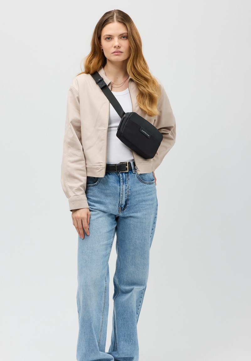 Schwarze Stoff-Schultertasche im Crossbody-Stil, rechteckige Form, mit Reißverschluss, verstellbarer Trageriemen. Modell in beiger Jacke und blauen Jeans.