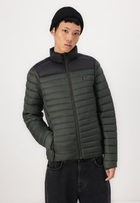 Grøn polstret jakke med en sort, glat øverste del. Har en ståkrave, lynlåsåbning og lodret syning. Båret over mørke jeans.