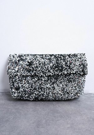 Kleine rechthoekige zilveren pailletten clutch met overslag, liggend op een grijze betonnen ondergrond tegen een witte achtergrond.