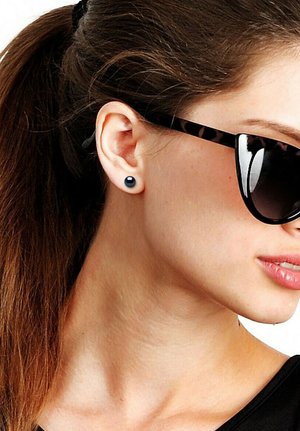 Femme aux cheveux bruns attachés en queue de cheval, portant des boucles d'oreilles rondes noires et des lunettes de soleil à monture écaille foncée, tournée vers la droite sur un fond blanc.