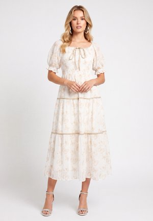 Femme portant une robe midi blanche à manches bouffantes, imprimé papillon discret, encolure carrée avec lien et talons argentés à bride cheville, debout devant un fond blanc.