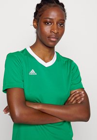 Zelený atletický dres vyrobený z hladkej tkaniny, so zloženým bielym V-krkom a malým bielym logom Adidas na hrudi.