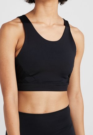 Femme portant un soutien-gorge de sport noir et des leggings noirs taille haute, montrée des épaules jusqu'au ventre sur un fond uni.