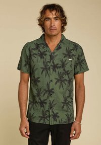 Chemise verte à manches courtes avec un motif de palmier, tissu texturé et une poche poitrine, portée avec un pantalon foncé.