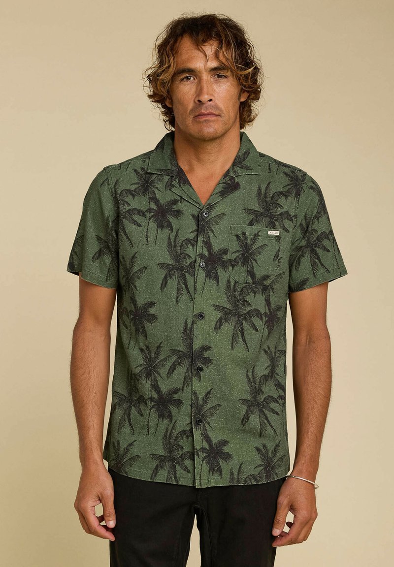 Chemise verte à manches courtes avec un motif de palmier, tissu texturé et une poche poitrine, portée avec un pantalon foncé.