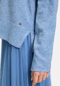 Hellblaues Sweatshirt aus strukturiertem Stoff, mit einem gerippten Saum und einem kleinen metallischen Akzent. Kombiniert mit einem plissierten blauen Rock.