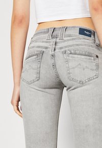 Woman wearing light gray fitted jeans with stitched back pockets and a white cropped top, shown from the waist down, back view.