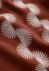 Tissu texturé marron avec des motifs brodés blancs en forme d’éventail répétés disposés en diagonale sur la surface.