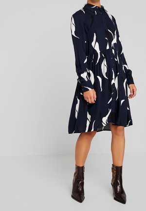 Kobieta ubrana w granatową sukienkę zapinaną na guziki z białymi abstrakcyjnymi wzorami oraz brązowe teksturowane botki za kostkę, stojąca z rękami swobodnie opuszczonymi wzdłuż ciała.