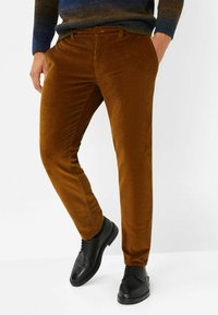 Braune Cordhosen im geraden Schnitt, mit feinen vertikalen Rippungen und Seitentaschen. Getragen mit schwarzen Schnürschuhen.