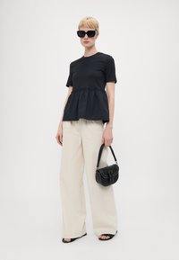 Svart kortermet topp med peplum-hem, kombinert med løse kremfargede bukser og en liten svart veske. Modellen har på seg store svarte solbriller.