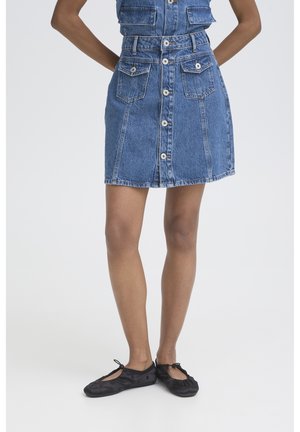 Jambes d'une personne portant une jupe en jean bleue à boutons sur le devant avec des poches et des ballerines noires sur fond blanc.