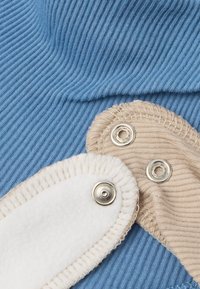 Tessuto a coste di colore blu chiaro con toppe accentate beige e bianche dotate di chiusure a pressione. Le aree testurizzate creano un contrasto visivo.
