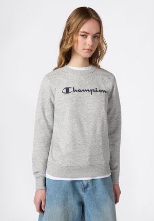 Junge Frau trägt ein hellgraues Champion Sweatshirt und hellblaue Jeans, steht vor einem schlichten weißen Hintergrund.