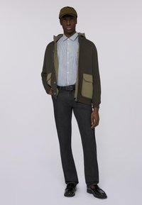 Cárdigan de punto suave en verde oliva con bolsillos grandes sobre una camisa de botones a rayas, combinado con jeans oscuros y mocasines negros, rematado con una gorra.