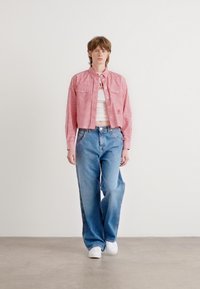Randig kort jacka i rött och vitt över en vit topp, ihop med vida blå denimjeans och vita sneakers. En enfärgad bakgrund.