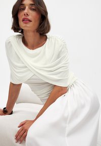 White dress featuring draped fabric at the shoulders, a fitted bodice, and a flowing skirt. Smooth texture with gathered detailing.
