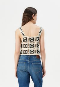 Top de tirantes recortado con un patrón floral de ganchillo en negro y crema, con tirantes anchos y un borde con forma de onda, combinado con unos jeans de cintura alta en azul.