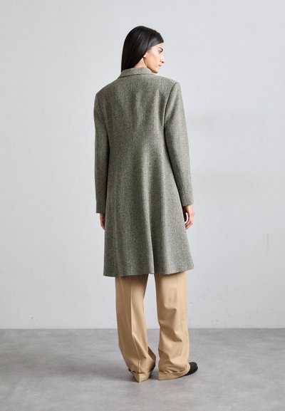 Herringbone-patterned, long grey coat with a collar, tailored silhouette, and long sleeves, paired with tan wide-leg trousers.