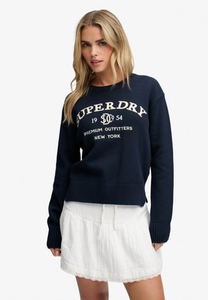 Lyshåret kvinde iført en navyblå sweater med hvid tekst og et kort hvidt skørt med blonderkant mod en ensfarvet baggrund.