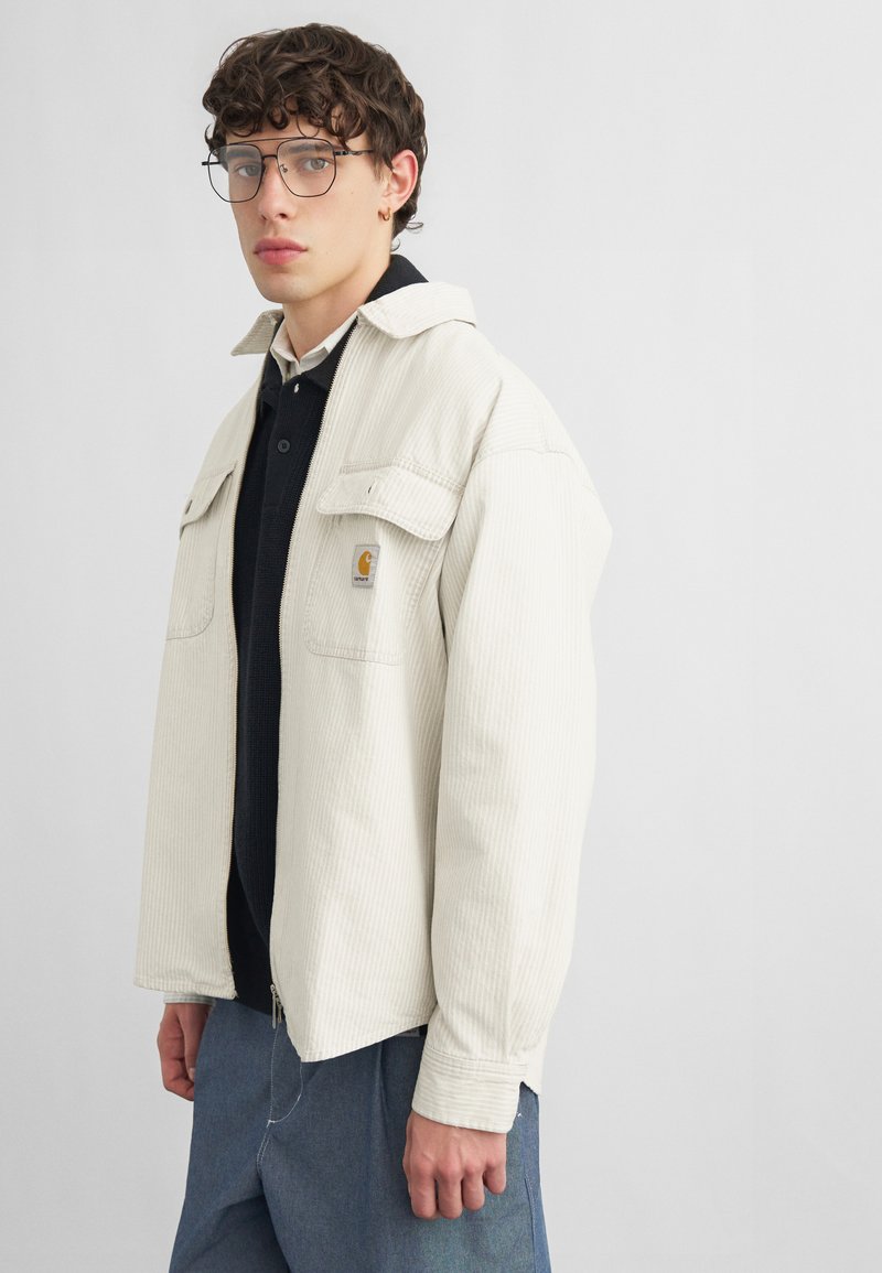 Junger Mann mit lockigem Haar und Brille, trägt eine cremefarbene Cordjacke über einem schwarzen Hemd und blauen Jeans, steht vor einem einfarbigen Hintergrund.