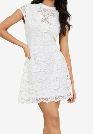 Robe blanche en dentelle à manches courtes avec motifs floraux et embellissements de fleurs en 3D, taille cintrée et ourlet festonné portée par une femme debout.