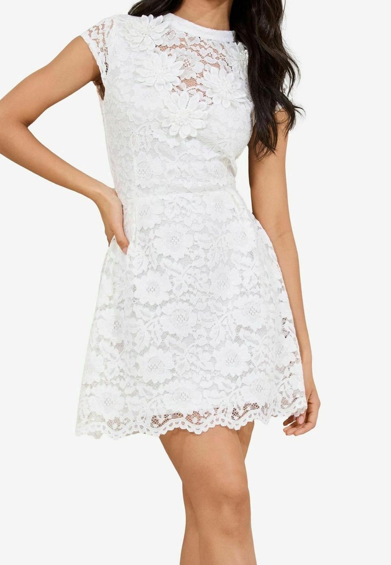 Robe blanche en dentelle à manches courtes avec motifs floraux et embellissements de fleurs en 3D, taille cintrée et ourlet festonné portée par une femme debout.