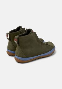 Las botas de tobillo verde oliva presentan una textura de ante suave, suelas azules y un cierre con cremallera. El logotipo de la marca es visible en los tacones.