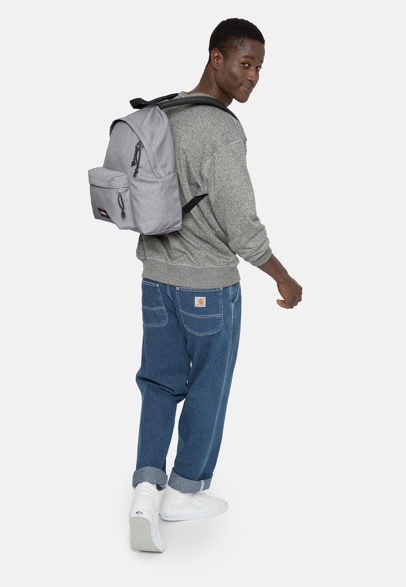 Grauer Stoffrucksack mit Fronttasche, Reißverschluss und verstellbaren Trägern. Von einer Person in lässiger Kleidung über der Schulter getragen.