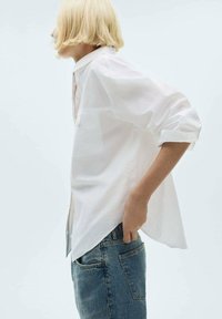Persona con capelli biondi corti indossa una camicia bianca con le maniche arrotolate e jeans blu, in piedi di lato con una mano in tasca.