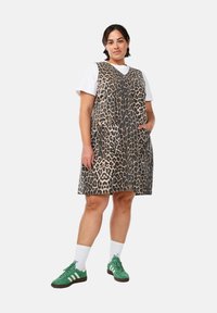 Leopardprint jurk met V-hals, mouwloos ontwerp, twee voorzakken en een losse pasvorm, gecombineerd met groene sneakers en witte sokken.