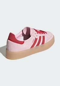 Roze suede sneaker met rode accenten, drie rode strepen aan de zijkant en een doorzichtige gumzool. Laag boven ontwerp met veters.