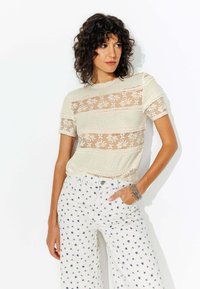 Haut à rayures en dentelle, à manches courtes, en matière beige clair. Associé à un pantalon large blanc avec de petits motifs floraux bleus.