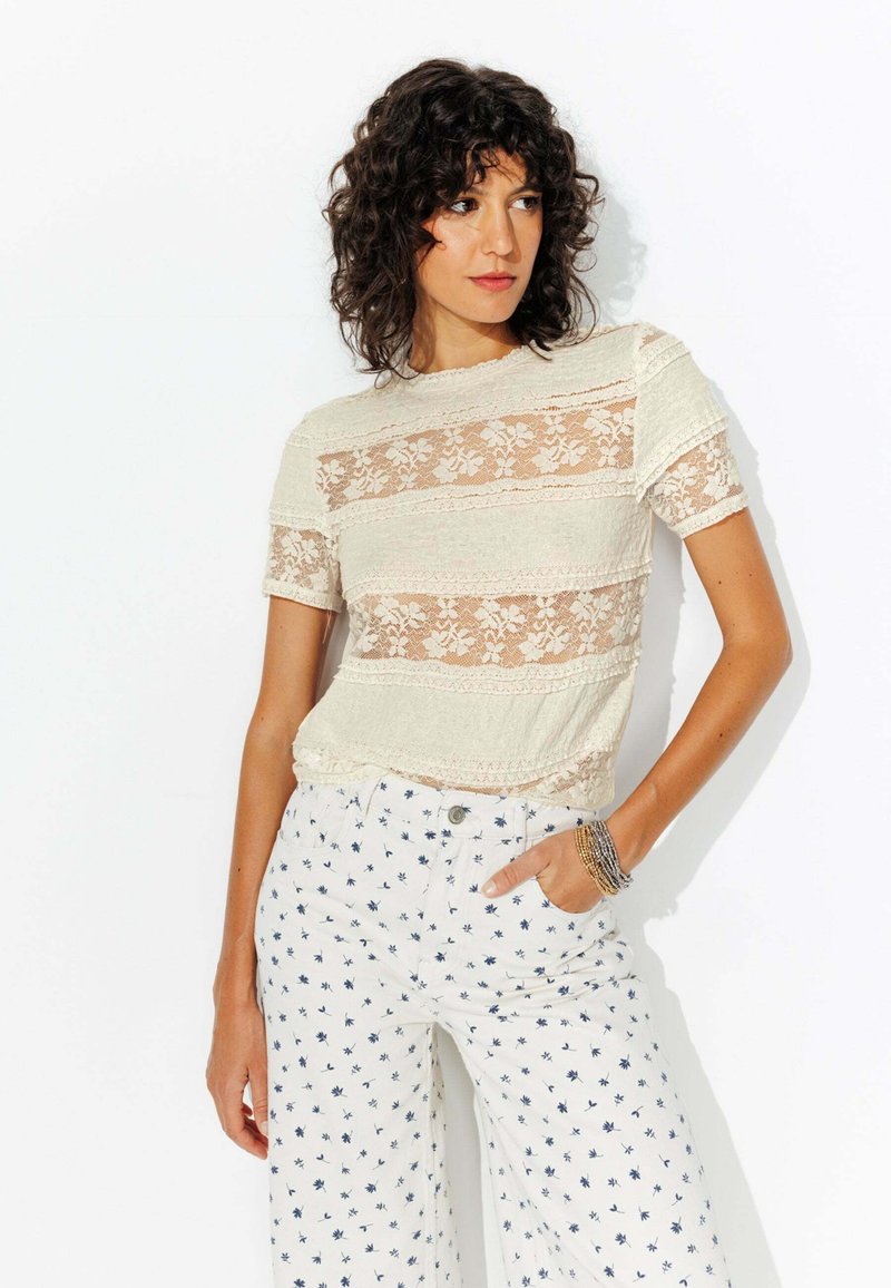 Haut à rayures en dentelle, à manches courtes, en matière beige clair. Associé à un pantalon large blanc avec de petits motifs floraux bleus.