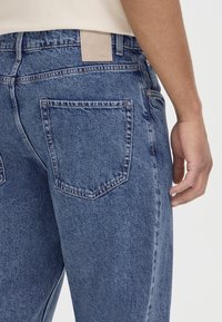 Gros plan d'une personne portant un jean en denim bleu avec une poche arrière visible, des passants de ceinture, et une partie d'une chemise beige.