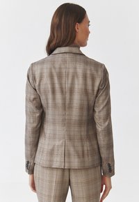 Hnědý kostkovaný blazer s přiléhavým střihem, který má klopový límec, zapínání na knoflíky na rukávech a strukturované švy na zadní straně.