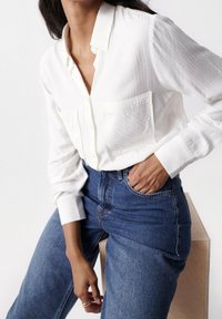 Femme portant une chemise blanche texturée boutonnée rentrée dans un jean bleu taille haute, assise sur une caisse en bois avec une main dans la poche.